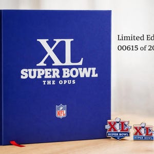 Op de afbeelding: Een blauw boek in beperkte oplage getiteld "XL Super Bowl The Opus" met witte letters en het NFL-logo. Twee bijpassende spelden worden naast het boek weergegeven. De tekst "Limited Edition 00615 of 20,000" is ook zichtbaar.