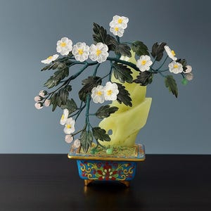 Op de afbeelding: Decoratieve bonsai-boomsculptuur met witte bloemen met gele centra en donkergroene bladeren. Het staat op een lichtgroene, jade-achtige basis in een blauw-gouden, sierlijke pot. De achtergrond is donkerblauw.