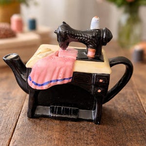 Puede incluir: Tetera negra y crema con forma de máquina de coser antigua. La tetera presenta un detalle de tela rosa con ribete azul, una máquina de coser negra y una bobina de hilo blanca. La tetera está sobre una superficie de madera.