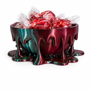 Puede incluir: Un cuenco decorativo con un diseño goteante, lleno de caramelos rojos envueltos. El cuenco tiene un acabado que cambia de color, pasando del verde azulado al rojo. Los caramelos tienen la palabra "BIRTHDAY" impresa.