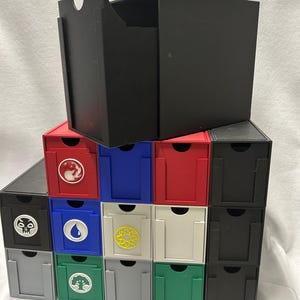 Puede incluir: Una pila de cajas de almacenamiento de cartas de colores. Las cajas son rojas, azules, blancas, verdes, grises y negras. Cada caja tiene un símbolo diferente en la parte delantera. Una gran caja negra encima.