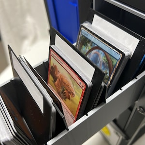 May include: A black plastic trading card storage box with multiple compartments containing stacks of collectible cards. The visible cards include an Ingot Chewer card with red artwork and an Island card with blue artwork, separated by black dividers.