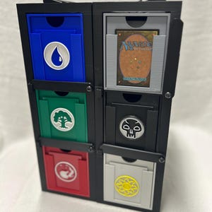 May include: A black, multi-compartment card storage unit with six drawers. Each drawer is a different color: blue, green, red, gray, and black. The drawers feature symbols from a trading card game, and one drawer contains a card.