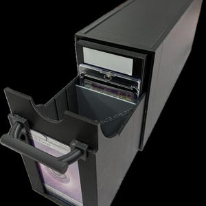 May include: A black card storage box with a sliding drawer. The drawer is open, revealing several trading cards in protective sleeves. The box has a handle and is designed to hold and organize collectible cards. The box is made of a solid, matte black material.
