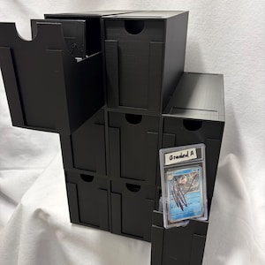 May include: A black modular storage unit with nine individual drawers, featuring a matte finish and circular finger pulls. One drawer is partially extended, revealing a clear plastic protective case containing a trading card labeled Graded A.