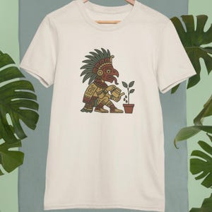 Puede incluir: Camiseta color crema con una ilustración detallada de una figura azteca regando una planta. La figura está representada en tonos marrones, verdes y rojos, con patrones intrincados. La planta está en una maceta marrón. La camiseta está colgada en una percha de madera.