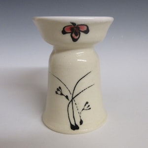 May include: A cream-colored ceramic candle holder with a flared base and a small, bowl-shaped top. The holder is decorated with a black floral design and a red and black flower on the top. The candle holder is approximately 4 inches tall.