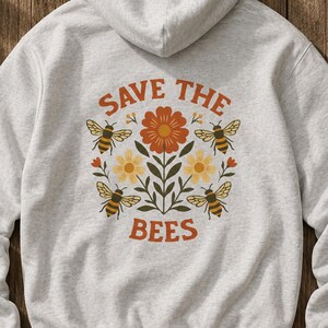 May include: A light gray hoodie with a floral and bee design. The design includes a large orange flower, yellow flowers, green leaves, and four bees. The text "SAVE THE BEES" is arched above and below the floral design.