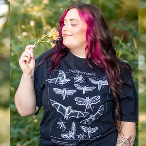 May include: A black short-sleeved t-shirt featuring a white graphic print of various nocturnal creatures labeled with letters and the text THE NIGHT SHIFT. The design includes illustrations of bats, moths, and insects arranged in a grid-like pattern.