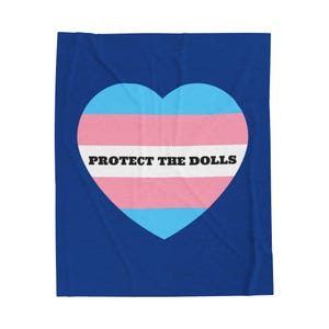 Könnte beinhalten: Eine blaue Decke mit einem herzförmigen Transgender-Pride-Flaggen-Design. Das Herz zeigt den Text "PROTECT THE DOLLS" in Schwarz.