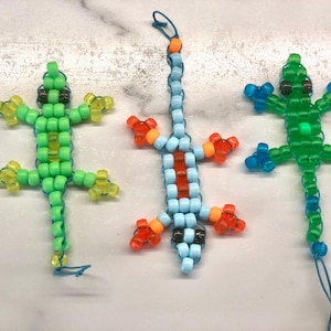 May include: Three beaded lizard ornaments in shades of green, blue, and orange. The lizards are made of small, colorful beads strung together to form the shape of a lizard. Each lizard has a loop for hanging.
