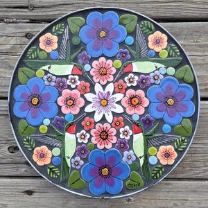 Könnte beinhalten: Runder, dekorativer Garten-Trittstein mit einem farbenfrohen Blumen- und Kolibri-Design. Der Stein hat einen dunkelgrauen Hintergrund und einen Metallrand. Die Blumen sind blau, rosa und orange, und die Kolibris sind grün und rot.
