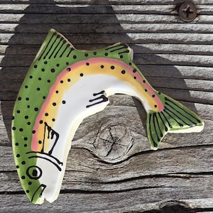 May include: A ceramic rainbow trout figurine with a green body, black spots, and a colorful stripe. The fish has a white belly and a detailed tail fin. The figurine is placed on a weathered wooden surface.