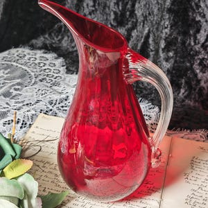 May include: A vibrant ruby red glass pitcher featuring a clear, ribbed glass handle and a curved pouring spout. The pitcher has a smooth, rounded body with vertical fluting that catches the light, creating a translucent and reflective appearance.