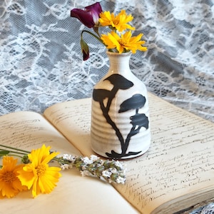 May include: A small, off-white ceramic vase with a ribbed texture and a black painted tree silhouette. The vase contains a single purple flower and two yellow flowers, with additional yellow flowers and lavender sprigs resting nearby.