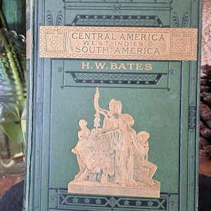 May include: An antique book titled "Central America, West Indies & South America" by H.W. Bates. The book has a green cover with gold lettering and a gold relief image. The book is displayed on a dark surface.