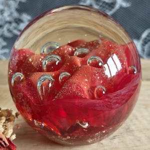 May include: A spherical glass paperweight featuring a deep red interior with layered petal-like forms and embedded gold-coloured flecks. The clear glass exterior contains several trapped air bubbles of varying sizes distributed throughout the upper section.