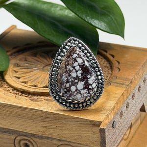 May include: A silver ring with a teardrop-shaped stone. The stone is a mix of brown and white, with a textured appearance. The ring is set on a wooden box with carved details, and green leaves are in the background.