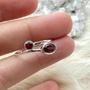 May include: A silver ring featuring two oval and round, deep red gemstones. The ring's band has a textured design. The gemstones are set in a silver setting. The ring is displayed on a finger.