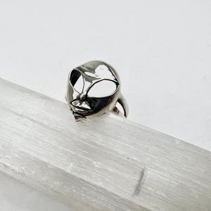 May include: A silver ring with an alien face design. The ring is sitting on a white surface.