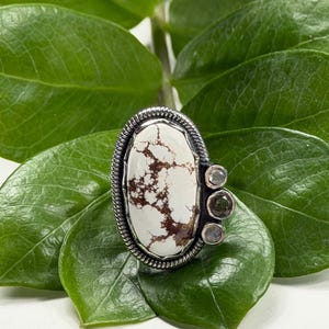 May include: Oval-shaped silver ring with a large white stone featuring brown veining. The ring has a textured silver border and is flanked by three smaller, round stones. The ring is set against a backdrop of green leaves.