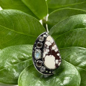 May include: A silver ring featuring a large, teardrop-shaped white stone with brown veining. The ring also has several smaller, round stones, including a moonstone. The ring is set against a backdrop of green leaves.
