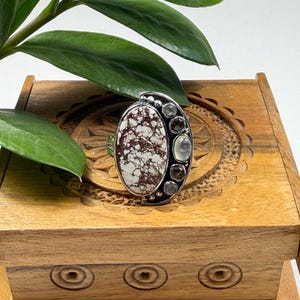 May include: A silver ring featuring an oval, white and brown patterned stone. The ring has a cluster of smaller, round stones in shades of gray and clear. The ring is displayed on a wooden box with carved details, with green leaves in the background.