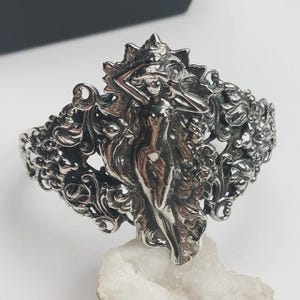 May include: A silver-toned cuff bracelet featuring a detailed, sculpted design. The bracelet showcases a central figure of a woman with flowing hair, surrounded by ornate floral and scrollwork patterns. The jewelry is displayed on a white, crystalline surface.
