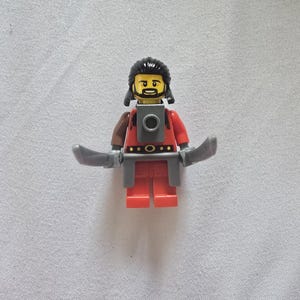 May include: A Lego minifigure of a warrior with black hair and a beard, wearing a red tunic and gray armor. The figure is holding two gray curved swords.