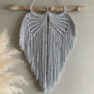 May include: A light grey macrame wall hanging, crafted by hand. The design is symmetrical, resembling wings or a leaf, and is suspended from a natural wooden branch. The macrame is made of soft, textured yarn, creating a bohemian aesthetic.