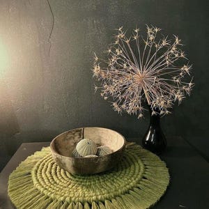 May include: A still life composition with a green fringed placemat, a wooden bowl holding sea urchins, and a dried flower arrangement in a black vase. The bowl sits on the placemat, creating a natural, earthy aesthetic.