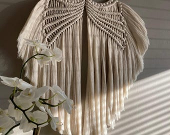 May include: Cream-colored macrame wall hanging with a textured design, suspended from a natural wooden branch. The intricate woven pattern features a central geometric design with flowing fringe. A decorative piece for home decor.