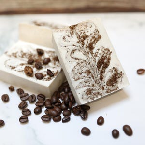 Könnte beinhalten: Drei rechteckige Seifenstücke mit Kaffeebohnenmuster und verstreuten Kaffeebohnen. Die Seife hat eine hellbeige Basis mit einem dunkelbraunen Wirbelmuster und Kaffeepulver darüber. Die Kaffeebohnen sind dunkelbraun.