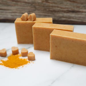May include: Three rectangular bars of light brown soap are arranged on a white marble surface, with a small pile of orange powder and several heart-shaped soap pieces. The background features a wooden texture.