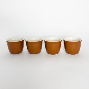May include: A set of four small, rounded cups with a smooth, glossy brown exterior finish and a contrasting white interior. Each cup features a simple, wide-rimmed design, uniform in shape and size, arranged in a horizontal row against a plain background.