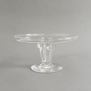 May include: A clear glass cake stand featuring a flat circular top plate supported by a central pedestal base. The pedestal has a decorative fluted design with vertical glass elements and rests on a stable, circular footed base.