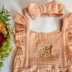 Könnte beinhalten: Ein babyfarbener Strampler mit Rüschen am Ausschnitt und Trägern. Der Strampler hat ein gesticktes Blumenmuster mit dem Buchstaben "M" in der Mitte. Ein passendes babyfarbenes Stirnband mit Schleife ist im Lieferumfang enthalten.