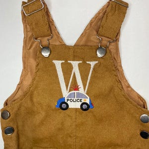 May include: Brown corduroy overalls with adjustable straps and metal hardware. The overalls feature a large white letter "W" and an embroidered police car with the word "POLICE" on the front. The overalls have two front pockets and snap closures.