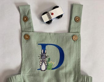 May include: A light green baby overall with a blue embroidered letter D and a rabbit illustration. The overall has wooden buttons on the straps.