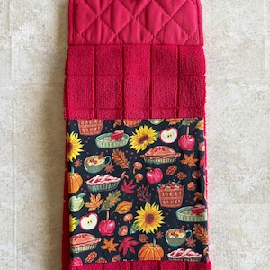 May include: A red hanging kitchen towel featuring a quilted top section with a wooden button closure and a lower section made of textured fabric. The center panel displays a black fabric print with sunflowers, pumpkins, apples, and pies.