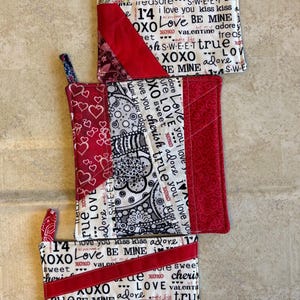 Puede incluir: Tres agarraderas de tela hechas a mano con varios patrones y texto. Las agarraderas presentan telas rojas, blancas, negras y verdes. El texto incluye "Te amo", "XOXO" y "Be Mine". Cada agarradera tiene un lazo para colgar.