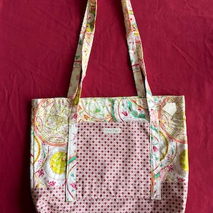 Puede incluir: Un bolso tote acolchado con un colorido estampado de cachemira y geométrico. El bolso presenta un patrón de diamantes rosa y blanco en la parte delantera, con una etiqueta "Princesses & Peas". Las asas y el borde superior tienen un diseño floral.