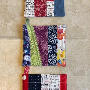 Puede incluir: Juego de tres agarraderas de tela hechas a mano. Una tiene un fondo azul con detalles de corazón rojo, otra presenta diseños florales y geométricos, y la última tiene un patrón de lunares rojos y blancos.