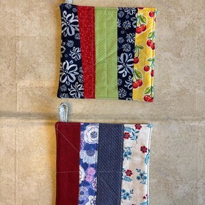 May include: Two square quilted pot holders. One features navy floral, red, green, and cherry print fabrics. The other has red, floral, blue, and cream floral fabrics. Each has a fabric loop for hanging.