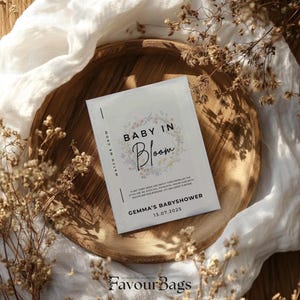 Pode incluir: Um cart&atilde;o branco com o texto "Baby in Bloom" em letras pretas com um design floral. O cart&atilde;o tamb&eacute;m inclui o texto "Gemma's Baby Shower" e a data "13.07.2025".