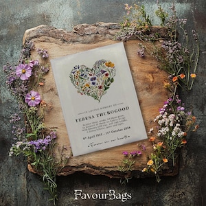 May include: A white card with a floral heart design and the text "In loving memory of Teresa Thurogood" with a poem about scattering seeds and a date range of April 1935 to October 2024. The card is on a wooden surface with purple flowers.
