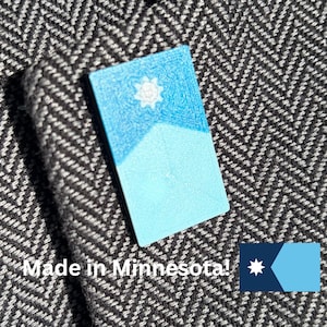 May include: A rectangular, blue and white painted wooden item with a sun and mountain design. The text "Made in Minnesota!" is below the item, along with a small flag of Minnesota.