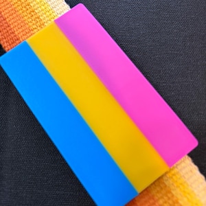 May include: A rectangular pin featuring the Pansexual Pride flag, with blue, yellow, and pink stripes. The pin is set against a black background and a strap with orange and yellow hues.