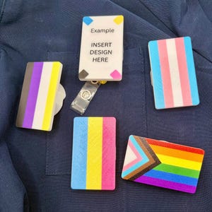 May include: Five rectangular badge reels with various flag designs. One white reel features the text Example INSERT DESIGN HERE with colorful corner accents. The others display non-binary, transgender, pansexual, and progress pride flag patterns.