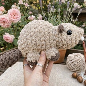 May include: A handmade crochet beaver in shades of beige and brown. The plush toy has a textured body, a flat tail, and small, dark eyes. The beaver is held in front of a blurred background of flowers and craft supplies.